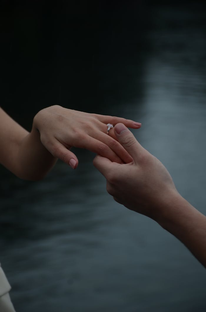 Intimate moment of exchanging an engagement ring outdoors by a serene water body.